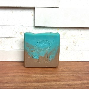 A rectangular bar of Earthy and citrusy soap, turquoise on top and brown on the bottom, embossed with a peacock feather and "Soaps by Glenda," displayed on a white tiled and wood background.